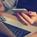 Person holding a smartphone near a laptop while managing business messages